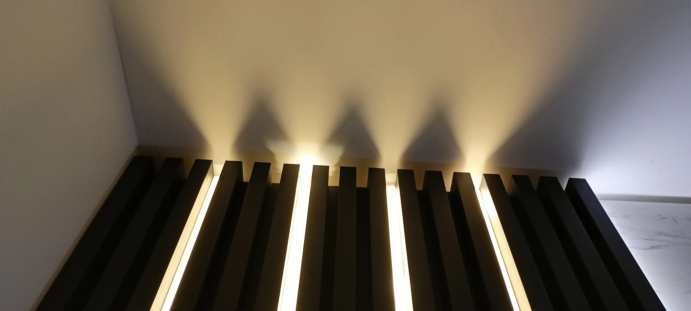 A wall of black vertical slats washed from above by warm light, each throwing its own shadow.